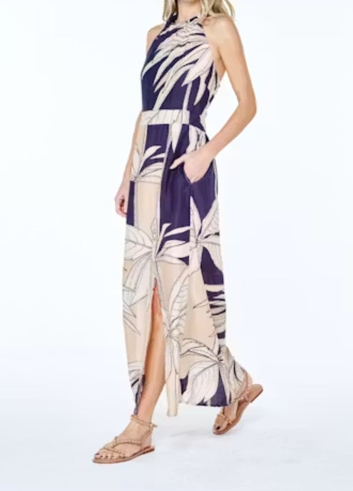A woman modeling a sleeveless halter-neck maxi dress with a tropical palm leaf print in navy blue and beige, paired with tan strappy flat sandals against a white studio background.