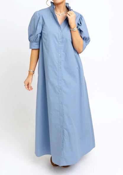 A woman wearing a light blue midi-length shirt dress with puff sleeves, a button-down front, and a ruffled collar, shown against a plain white studio background.