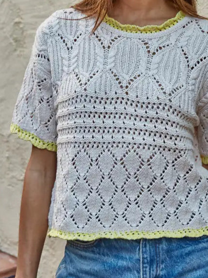 Woman wearing a light gray crochet top with green trim against a beige wall.