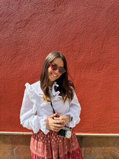 Woman in a white blouse and red patterned skirt standing against a red wall.
