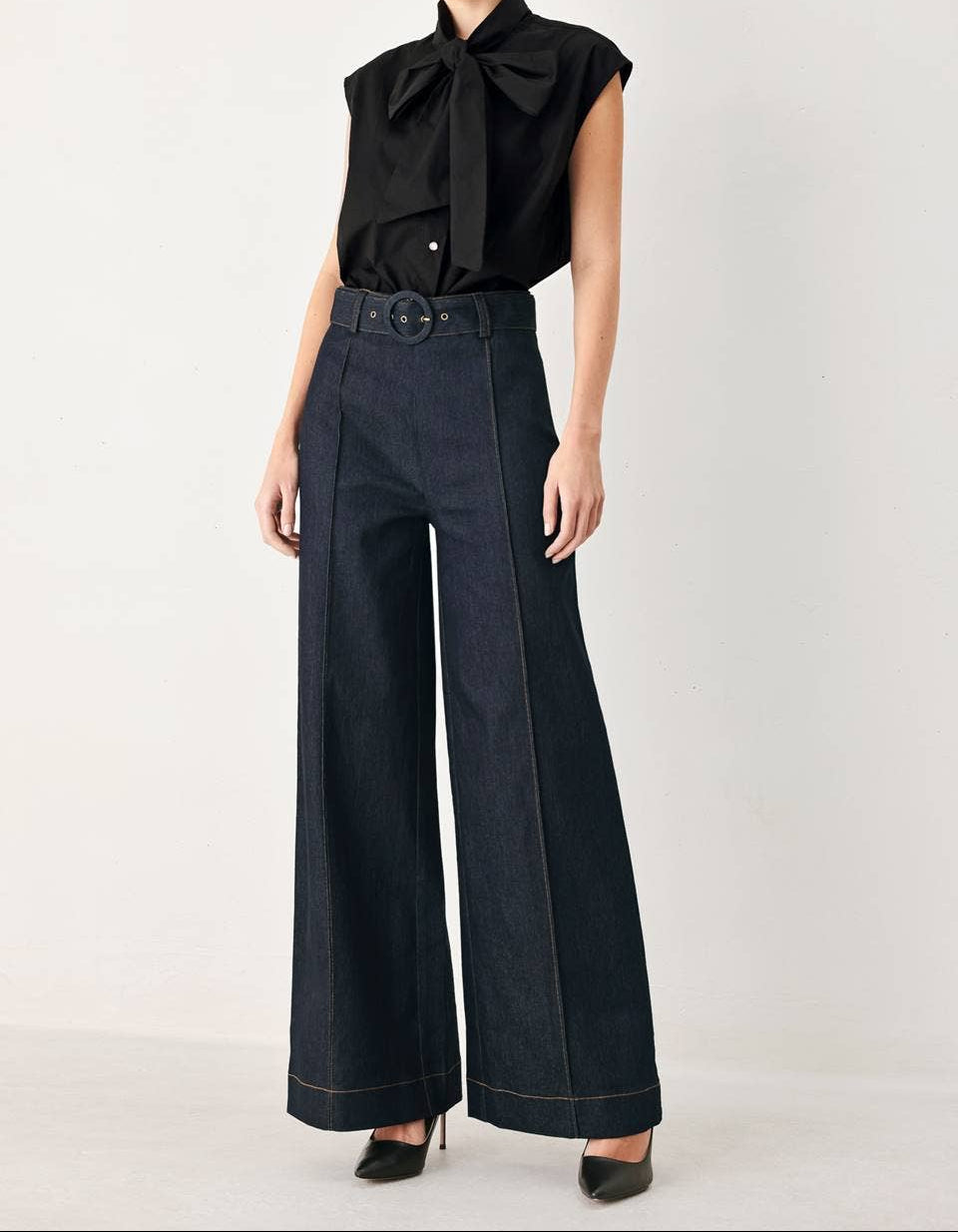 A model wearing a black sleeveless blouse with a large bow collar and dark wide-leg denim pants with a matching belt, standing against a plain light gray studio background.