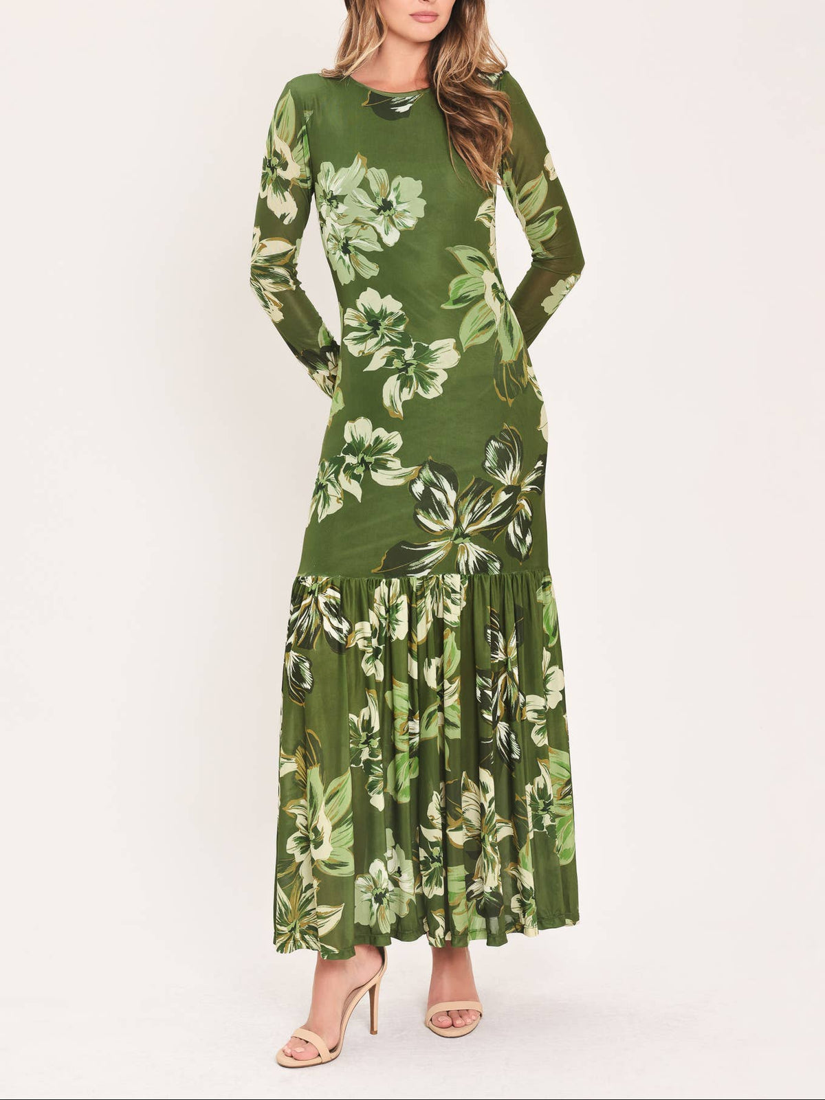 A woman modeling a long-sleeve green maxi dress with a large white and light green floral print, paired with beige high-heeled sandals, against a plain off-white studio background.