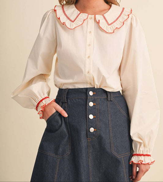 A woman wearing a cream-colored blouse with a large ruffled collar and cuffs trimmed in red stitching, paired with a dark blue denim skirt, against a solid beige background.
