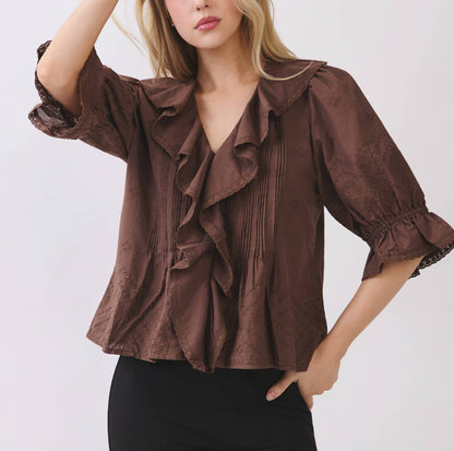 A woman modeling a brown ruffled blouse with three-quarter sleeves and black pants, posing against a light gray studio background.