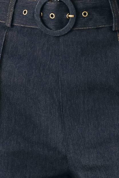 Close-up of dark indigo denim pants showing the waistband with a large circular ring detail and gold-tone metal eyelets.
