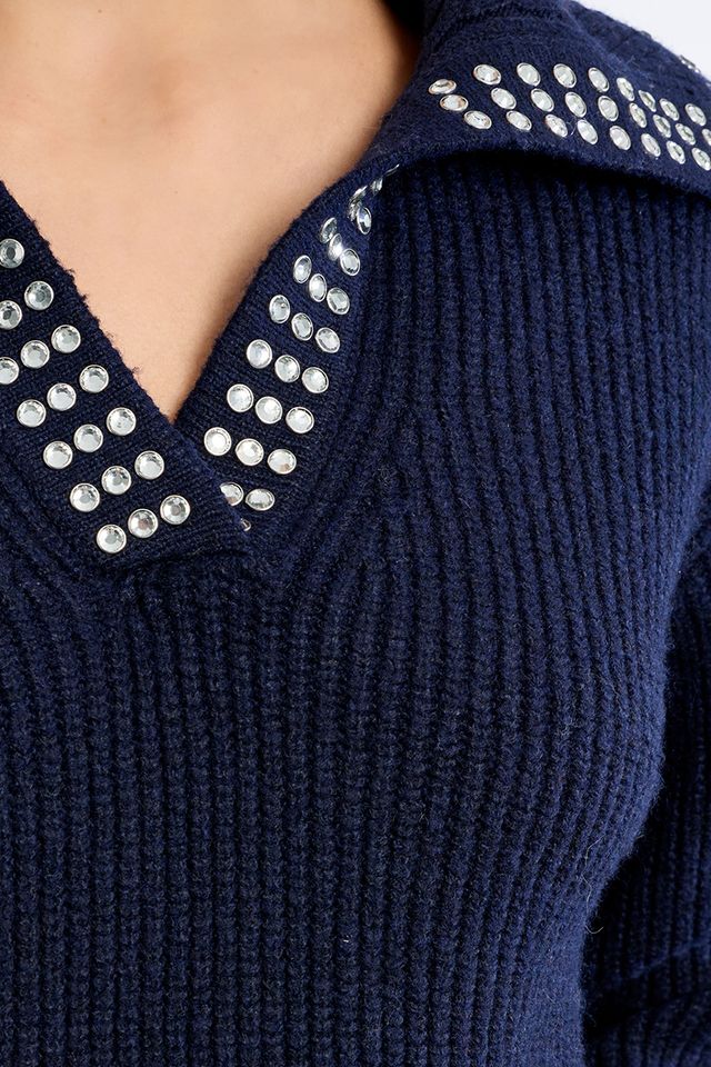 Close-up of a navy blue ribbed knit sweater with silver rhinestone embellishments around a V-neckline, worn by a model.