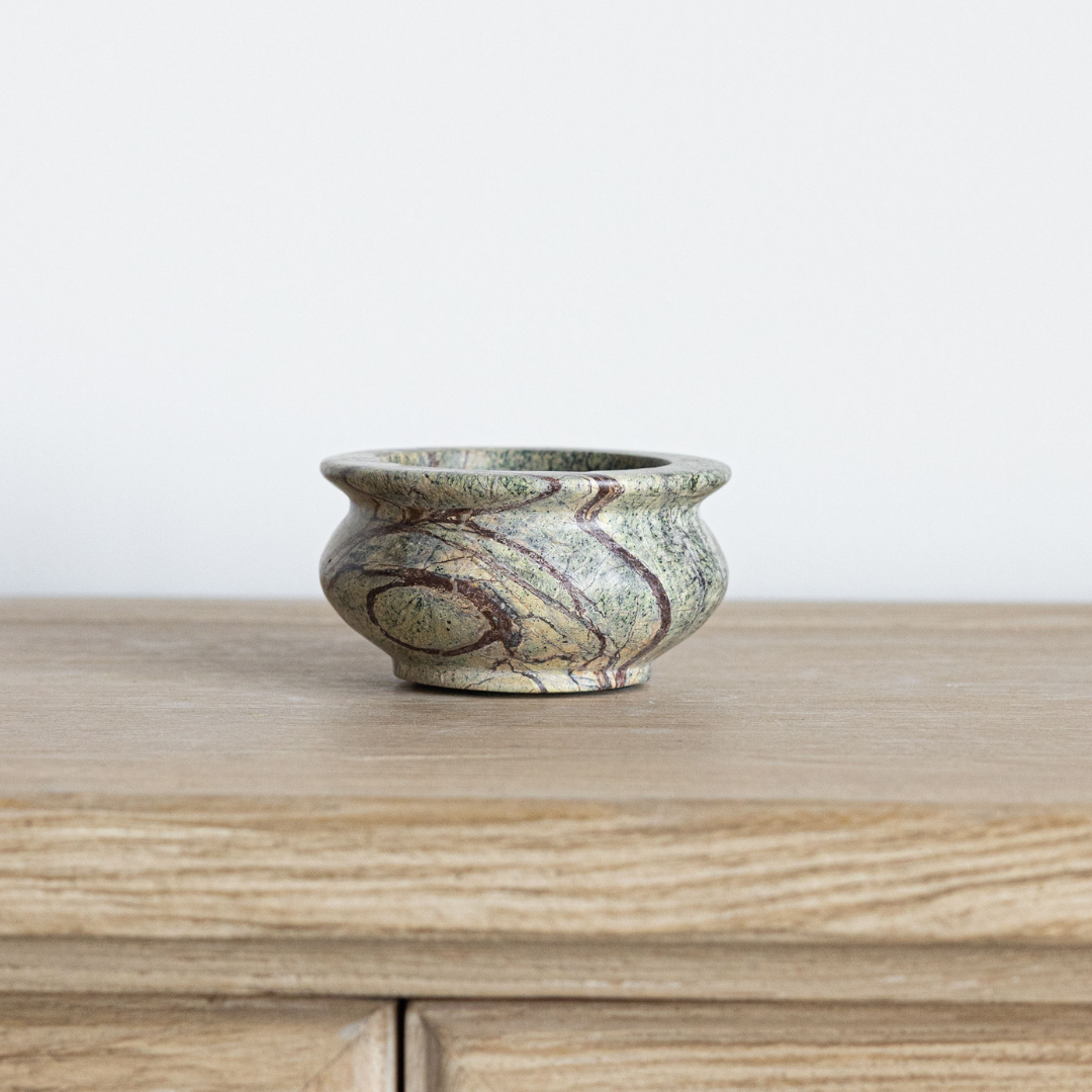 A small round stone bowl with a marbled pattern of green, beige, and brown, sitting on a light wood surface against a plain white wall.
