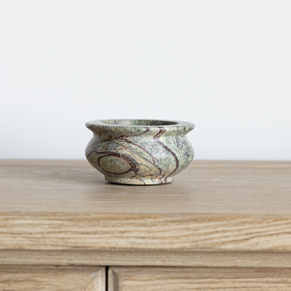 A small round stone bowl with a marbled pattern of green, beige, and brown, sitting on a light wood surface against a plain white wall.