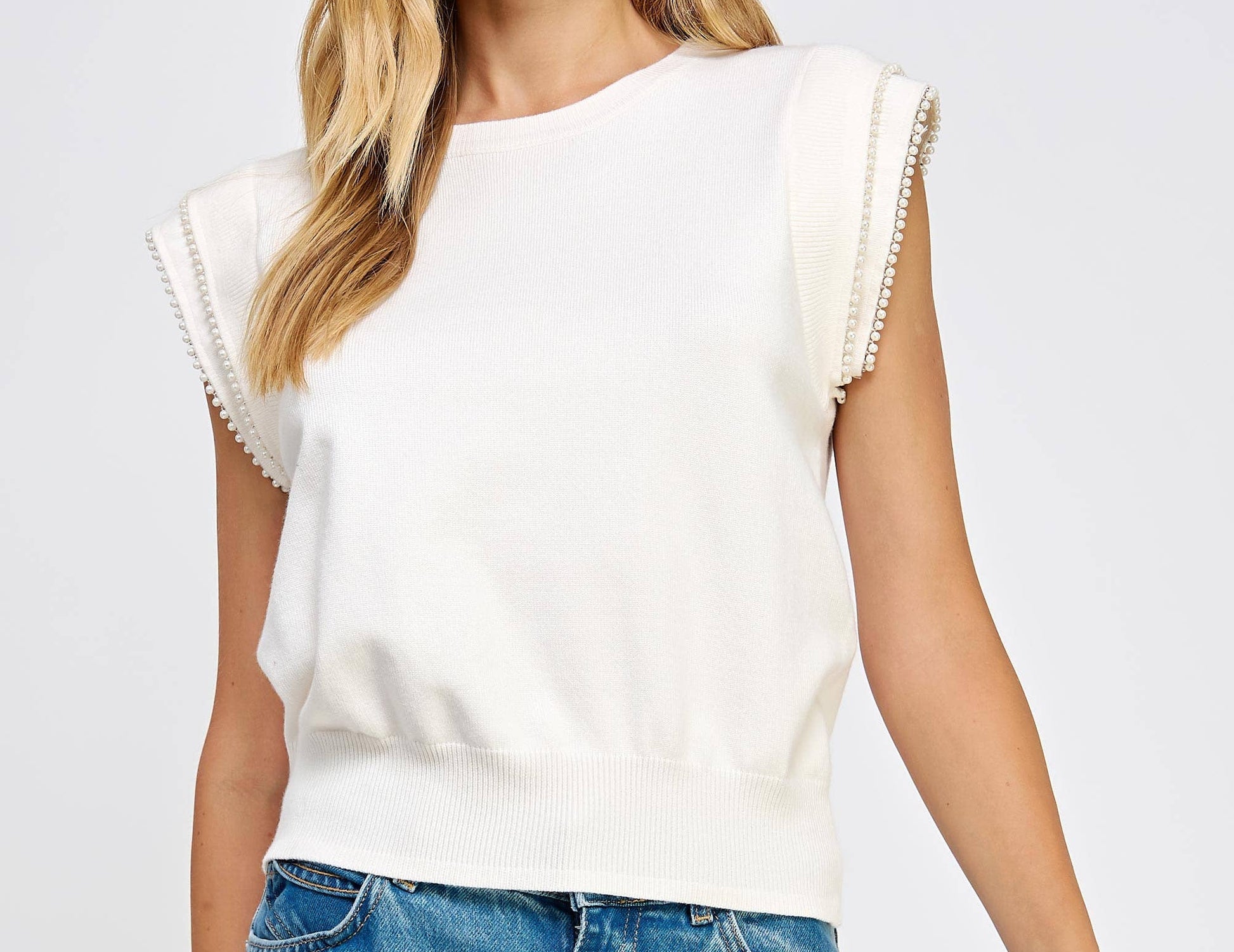 Woman wearing a white sleeveless top with ruffled sleeves and blue jeans on a light gray background