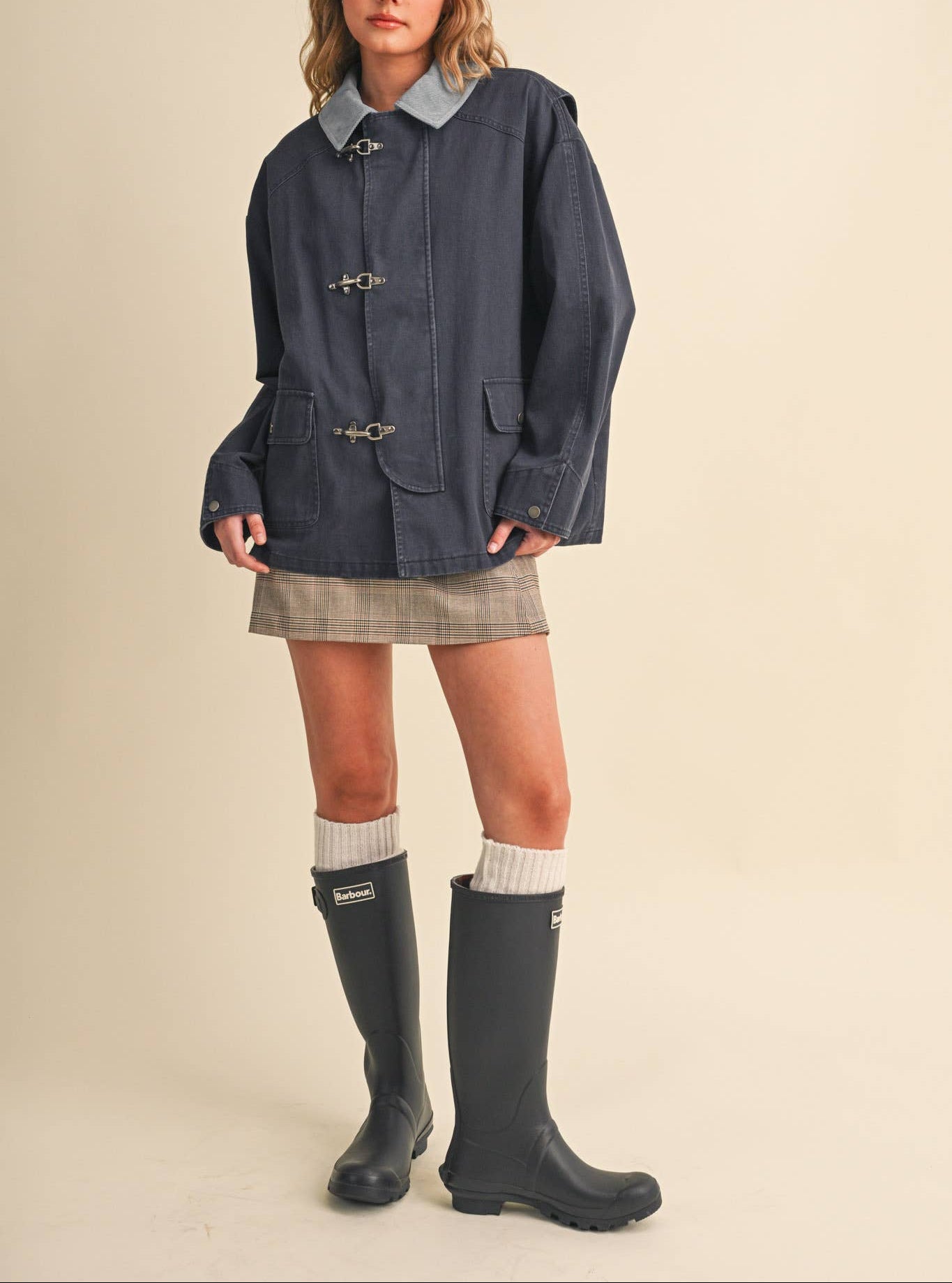 A woman modeling a dark navy blue waxed cotton jacket with toggle closures and a light blue collar, styled with a plaid mini skirt, white socks, and black rain boots against a solid beige background.