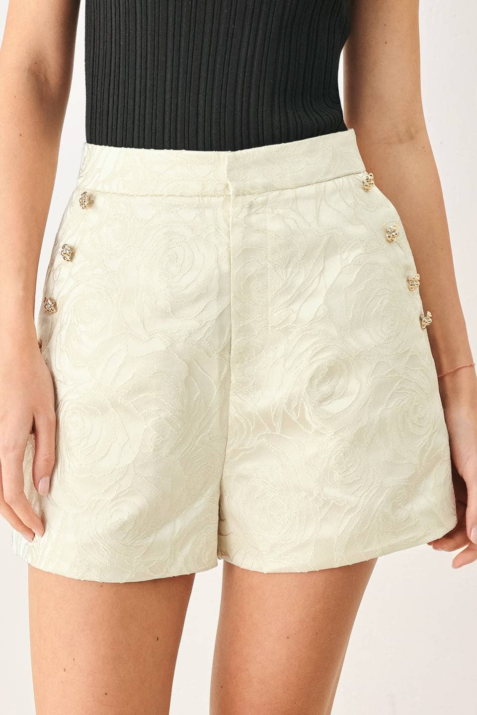A close-up of a model wearing high-waisted cream shorts with a subtle embossed swirl pattern and decorative gold-tone buttons down the sides, paired with a black ribbed top.