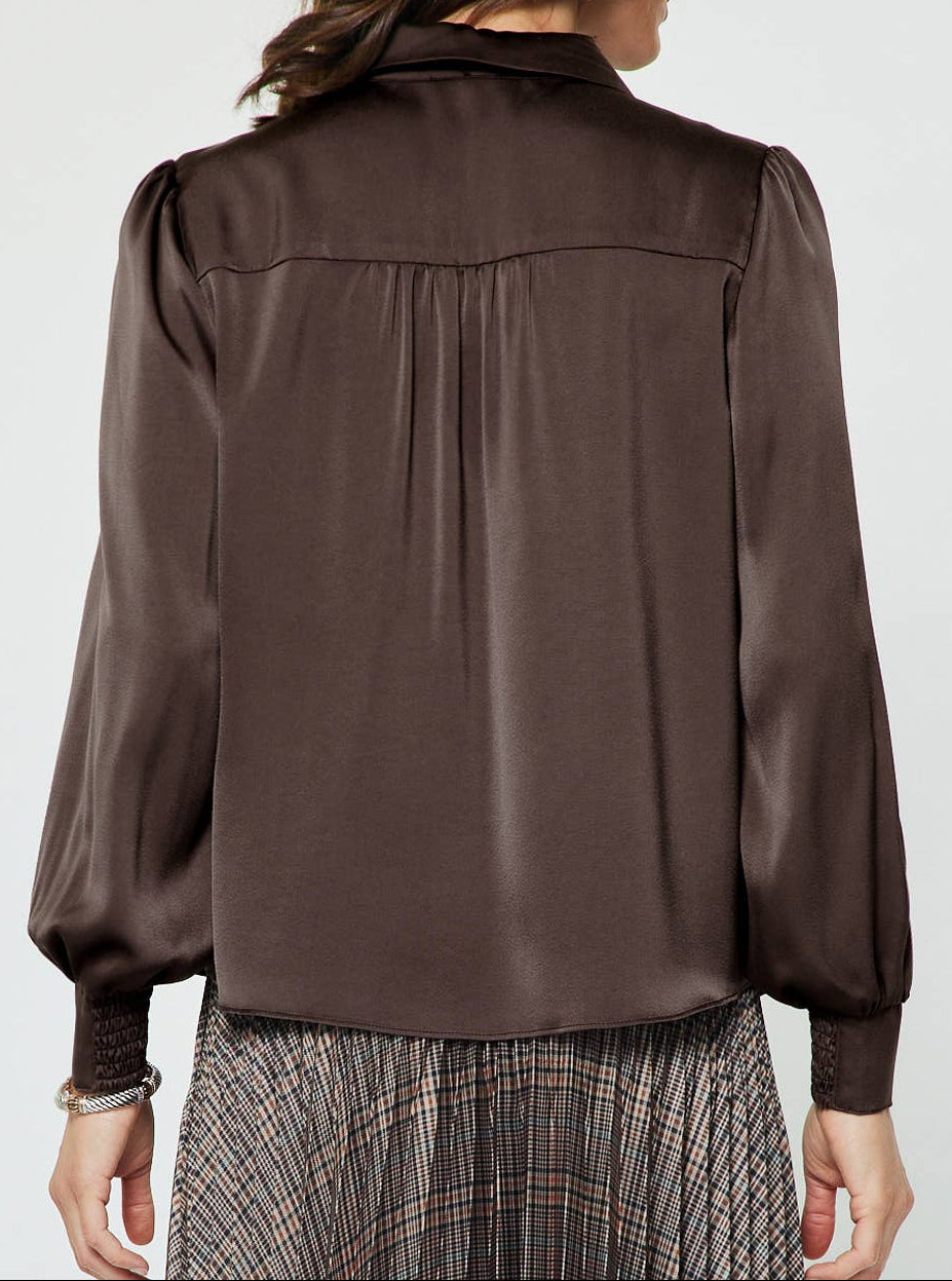 A woman viewed from the back wearing a dark brown satin blouse with long balloon sleeves and a gathered neckline, paired with a plaid skirt, against a light gray studio background.