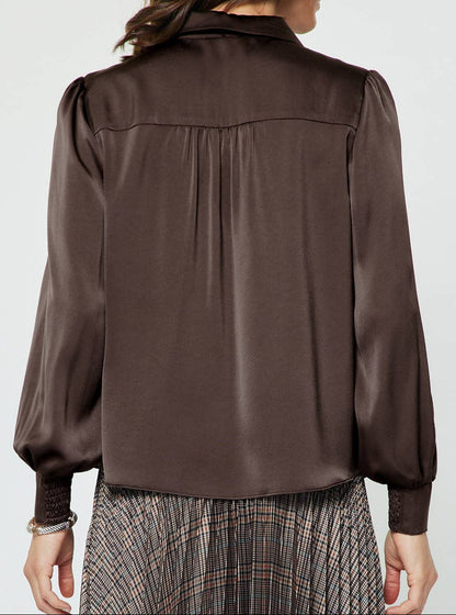 A woman viewed from the back wearing a dark brown satin blouse with long balloon sleeves and a gathered neckline, paired with a plaid skirt, against a light gray studio background.