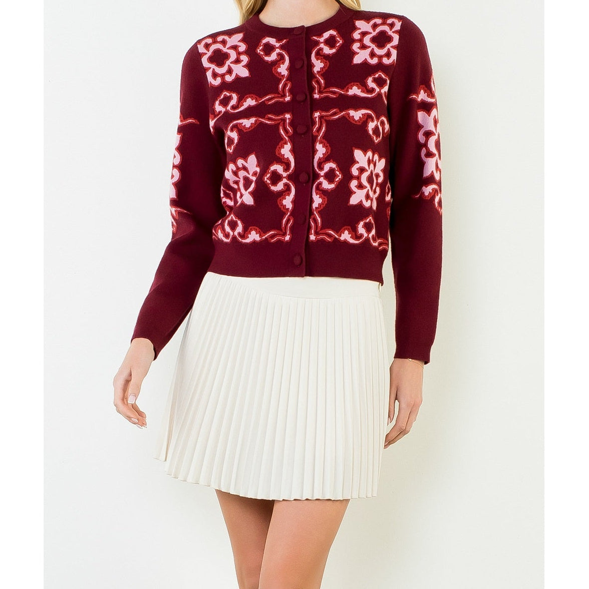 A woman modeling a burgundy cardigan with pink and white floral patterns, paired with a white pleated skirt, against a plain white studio background.
