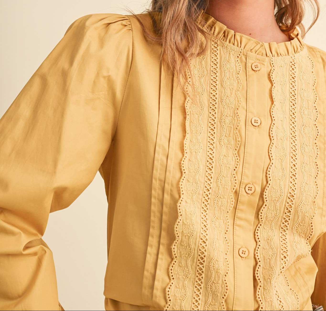 A woman wearing a pale yellow blouse with ruffled trim, vertical pleats, and lace-like front detailing, photographed against a plain warm beige background.