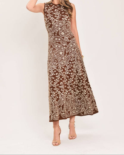 A woman modeling a sleeveless brown maxi dress with an intricate beige paisley pattern, standing against a plain light beige studio background.