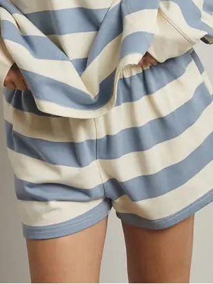 A woman wearing a matching two-piece loungewear set with horizontal light blue and cream stripes, featuring a long-sleeve half-zip pullover and matching shorts, set against a plain light gray studio background.