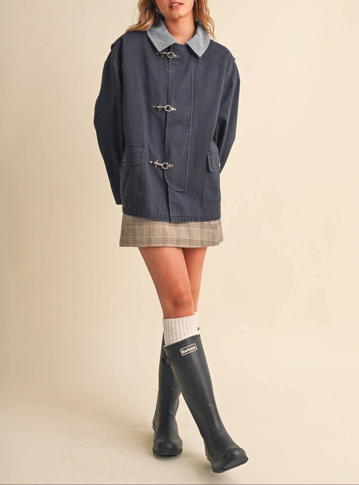 A woman modeling a dark navy blue hooded jacket with toggle closures, a light blue collar, a plaid mini skirt, white socks, and black rain boots against a plain beige studio background.