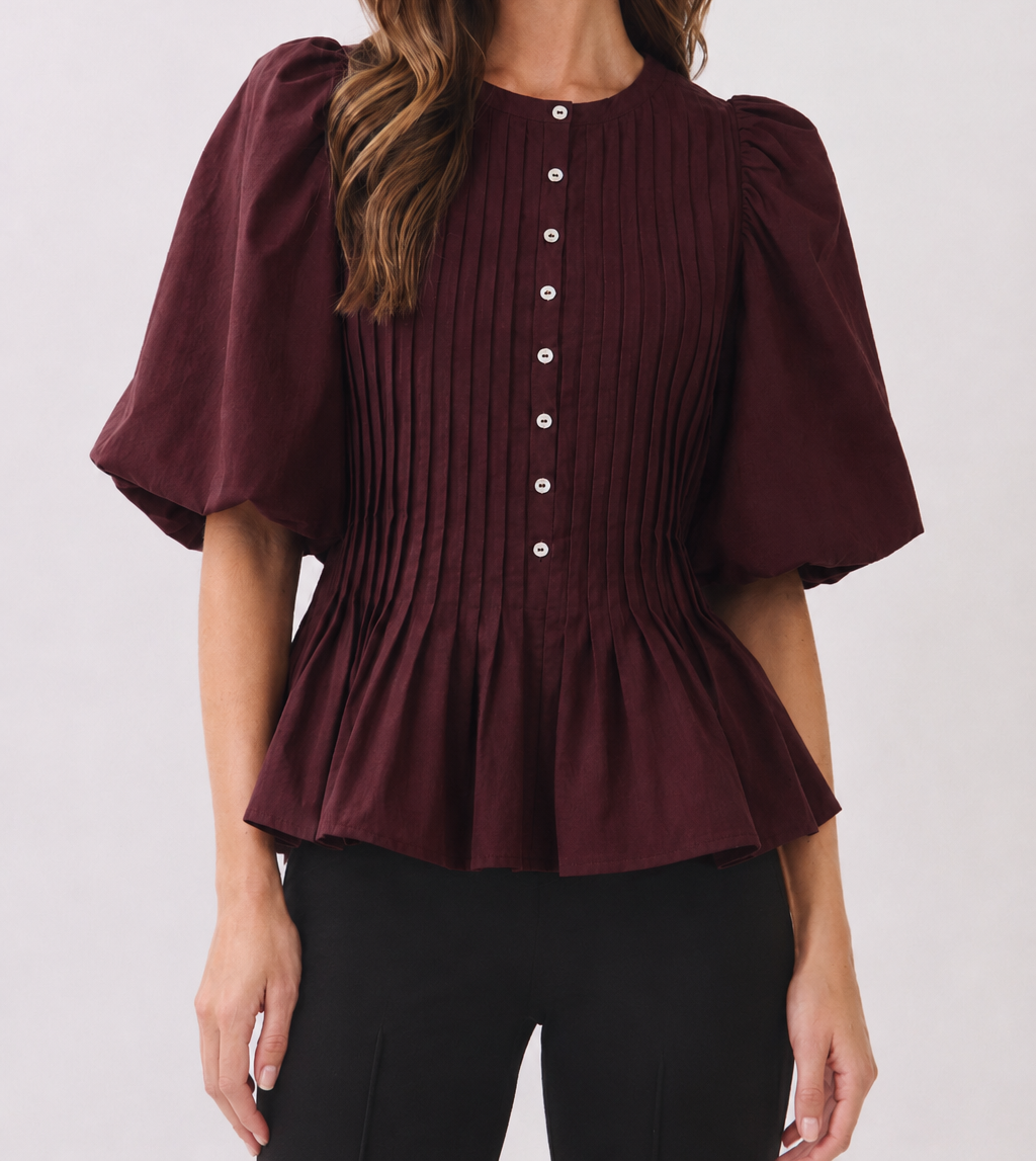 A woman wearing a burgundy short-sleeve blouse with puff sleeves, vertical pleating, and a button-down front, paired with black pants, against a light gray studio background.