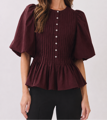 A woman wearing a burgundy short-sleeve blouse with puff sleeves, vertical pleating, and a button-down front, paired with black pants, against a light gray studio background.