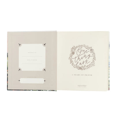 An open book or journal shown against a white background, with the left page in a light warm gray-beige tone and the right page in cream featuring a floral wreath and the words 'One thing at a time' and '3 Years of Prayer.'.
