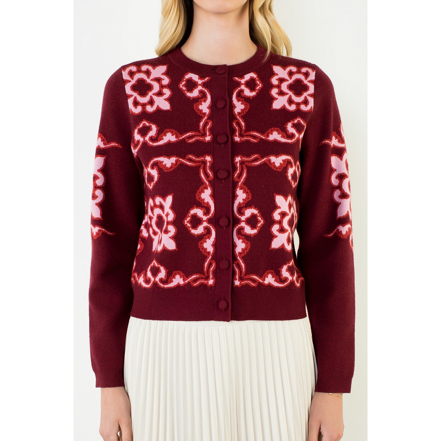 A woman wearing a burgundy cardigan with pink floral and scroll patterns, paired with a cream pleated skirt, shown against a plain light background.