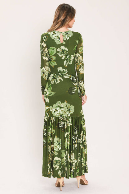 A woman viewed from the back wearing a long-sleeve olive green maxi dress with a large white and light green floral print, paired with beige high heels, against a plain off-white studio background.