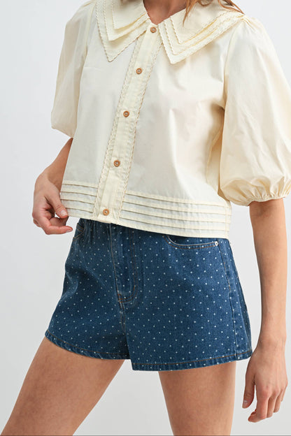 A woman modeling a cream-colored short-sleeve button-up blouse with a large decorative collar and puff sleeves, paired with blue patterned shorts, against a light gray studio background.