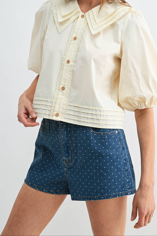 A woman modeling a cream-colored short-sleeve button-up blouse with a large decorative collar and puff sleeves, paired with blue patterned shorts, against a light gray studio background.
