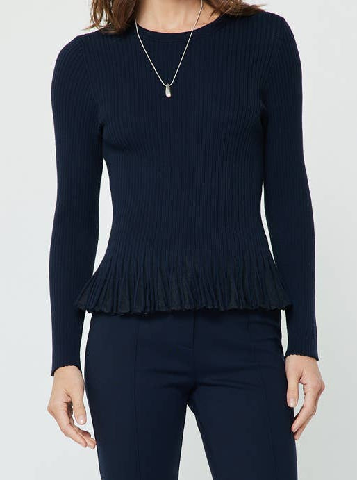 A woman modeling a dark navy blue knit long-sleeve peplum top and flared pants, paired with dark brown pointed-toe heels against a plain light gray studio background.