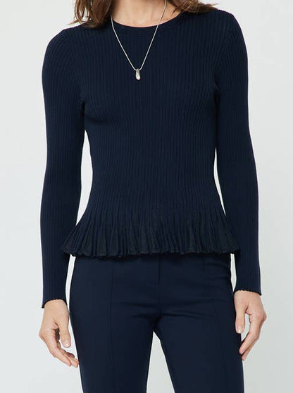 A woman modeling a dark navy blue knit long-sleeve peplum top and flared pants, paired with dark brown pointed-toe heels against a plain light gray studio background.