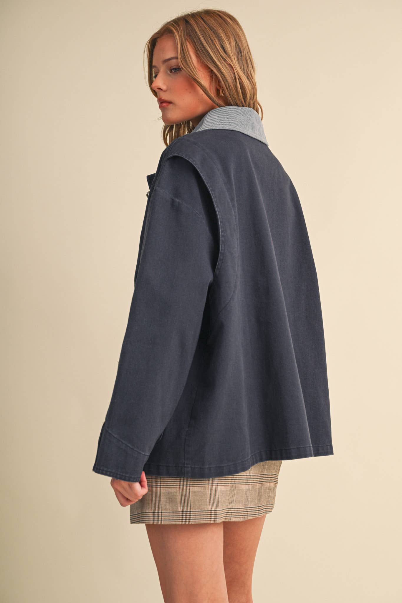 A woman shown from the back wearing an oversized navy blue utility-style jacket with a light blue collar over a beige plaid skirt, set against a plain beige studio background.