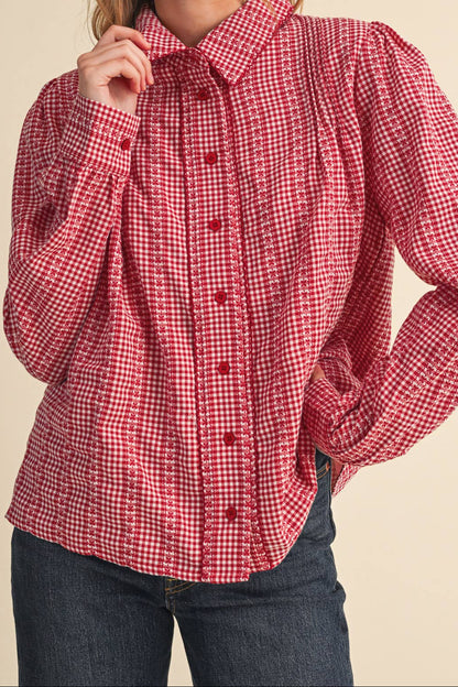 A woman wearing a red and white gingham long-sleeve blouse with a pleated front and button-down collar, paired with dark blue jeans, against a solid beige background.