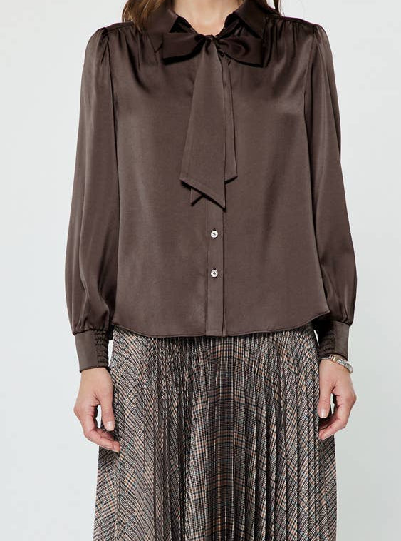 A woman modeling a dark brown long-sleeve satin blouse with a large bow at the collar and a matching pleated skirt, paired with dark brown ankle boots, against a plain light gray background.