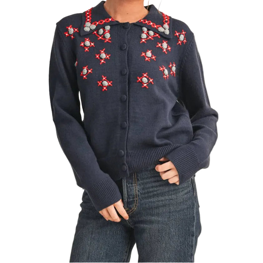 A woman modeling a navy blue button-up shirt with red and white floral embroidery on the chest, paired with dark blue jeans and red flats, against a solid beige studio background.