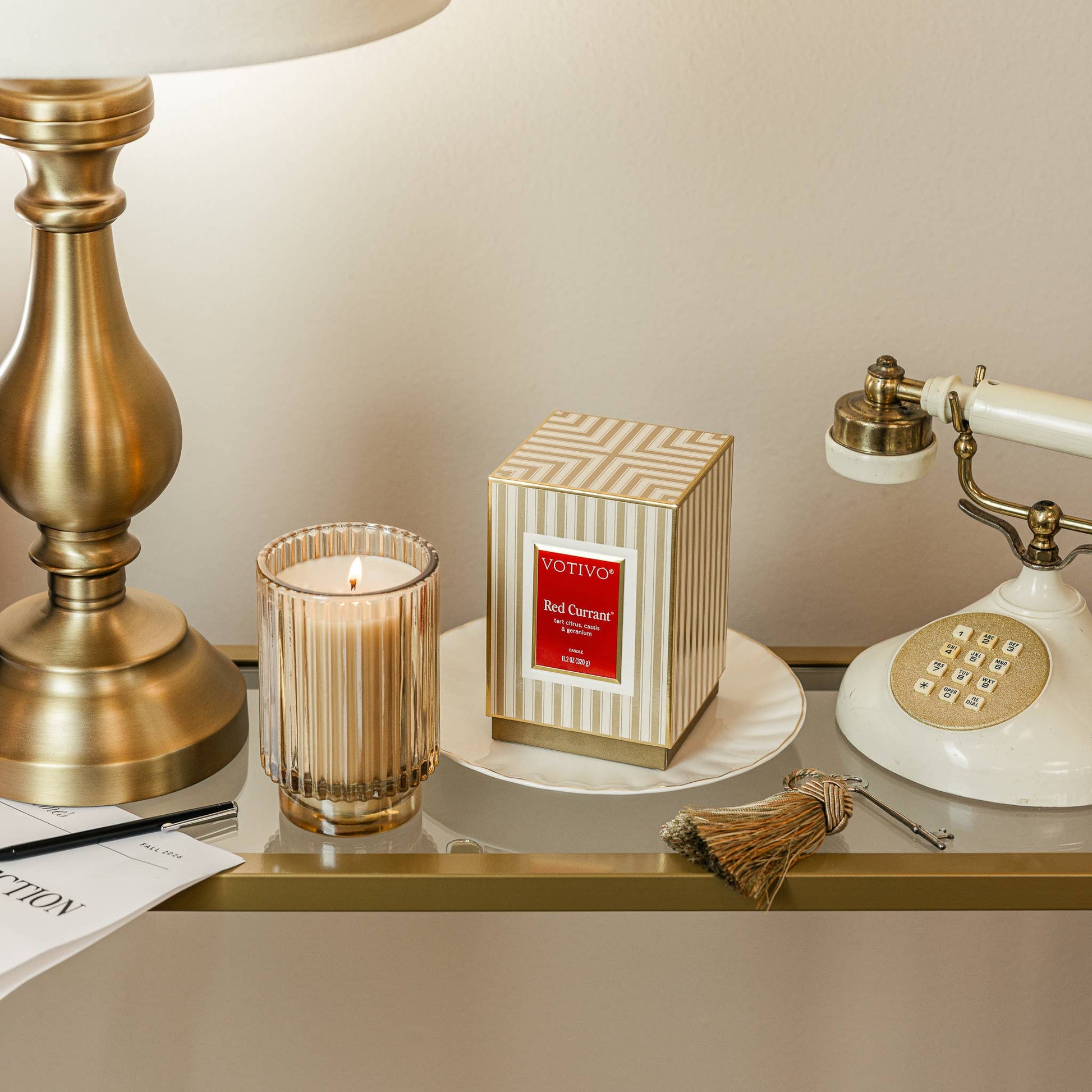 A VOTIVY 'Red Carpet' scented candle in a cream and gold striped box, styled on a shelf next to a lit striped glass candle holder, a brass table lamp, and a vintage cream rotary telephone.