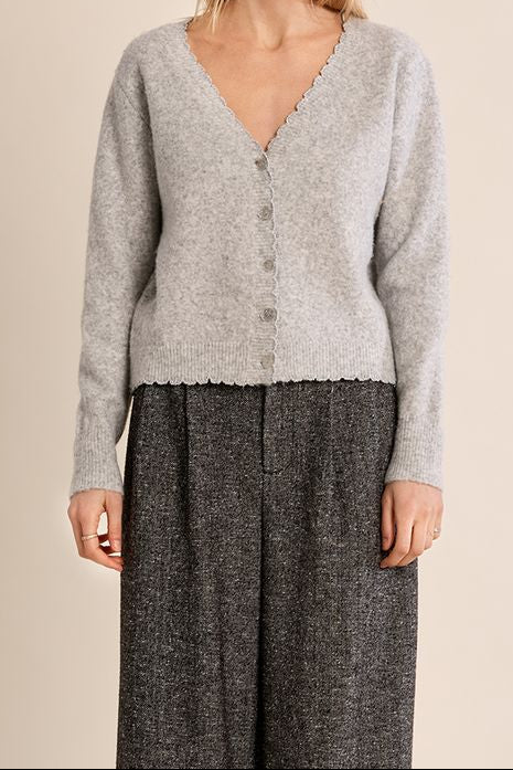 A woman modeling a light gray knit cardigan with a scalloped V-neck and button-front closure, paired with dark gray wide-leg trousers, against a plain beige studio background.