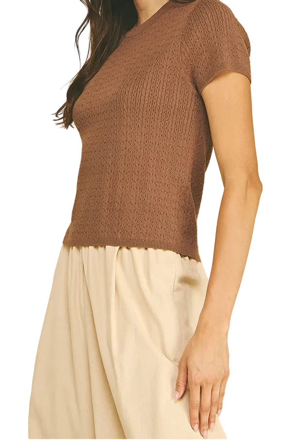 A woman wearing a brown short-sleeve ribbed knit top and cream-colored pants, standing against a solid beige background.