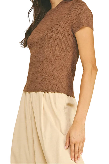 A woman wearing a brown short-sleeve ribbed knit top and cream-colored pants, standing against a solid beige background.