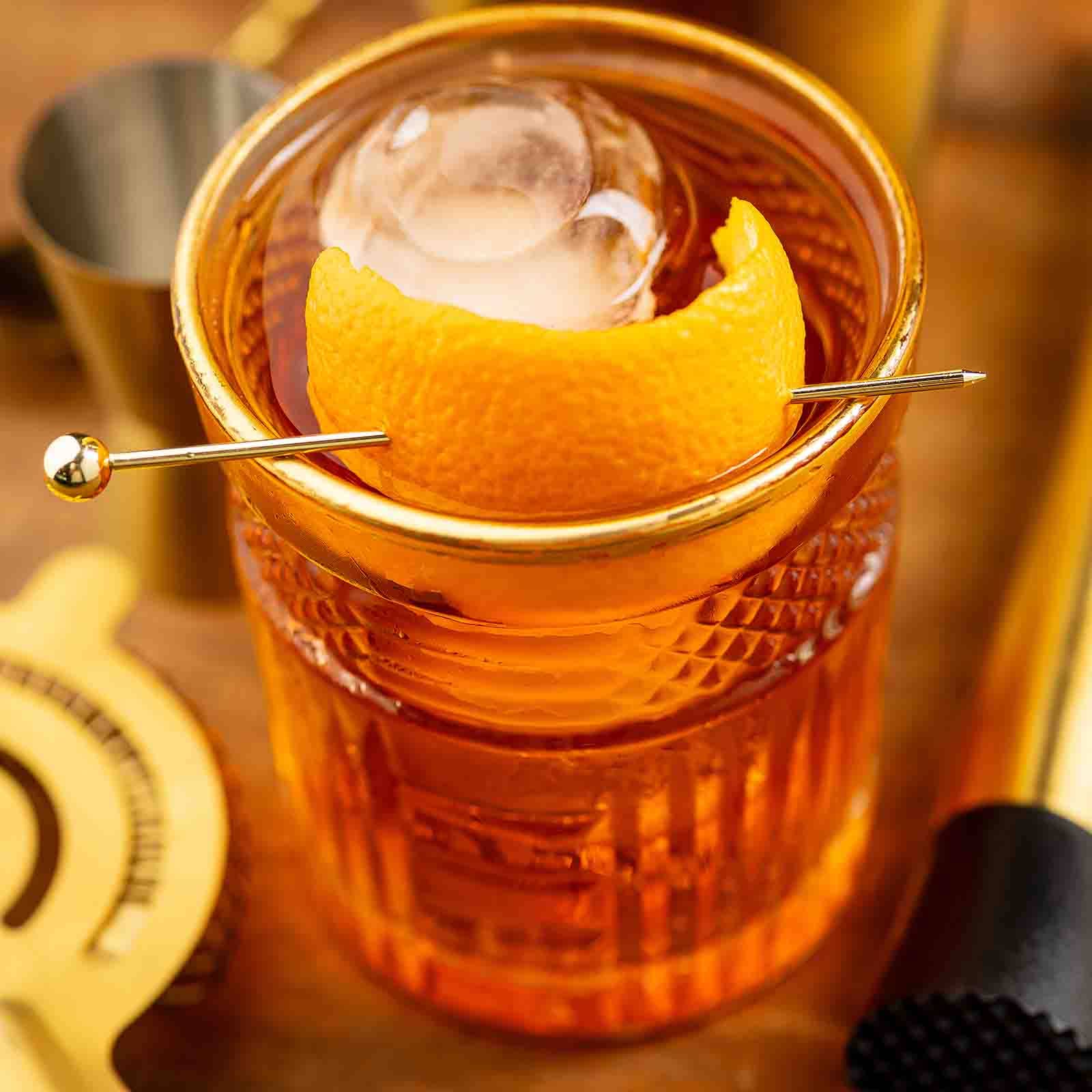 Cocktail with an orange twist and ice cube in a glass, garnished with a lemon peel, on a wooden surface.