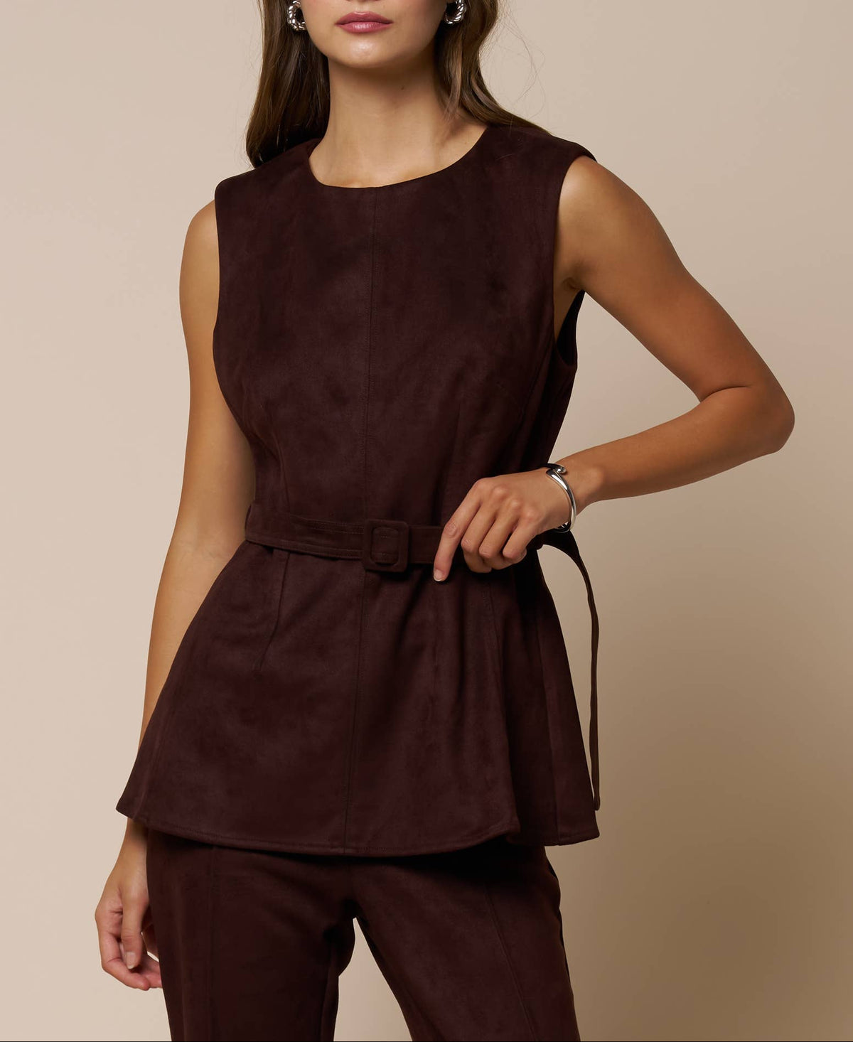 A woman modeling a sleeveless dark brown peplum top with a matching tie belt and matching dark brown trousers, set against a solid beige background.