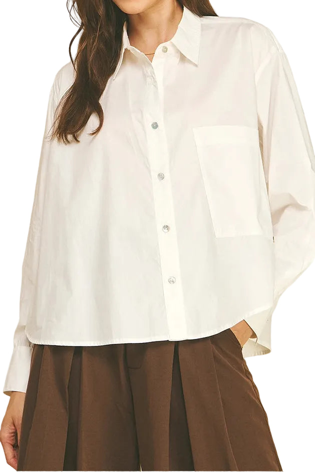 A woman modeling a white long-sleeve button-up shirt with a chest pocket, paired with brown pants, against a solid beige background.