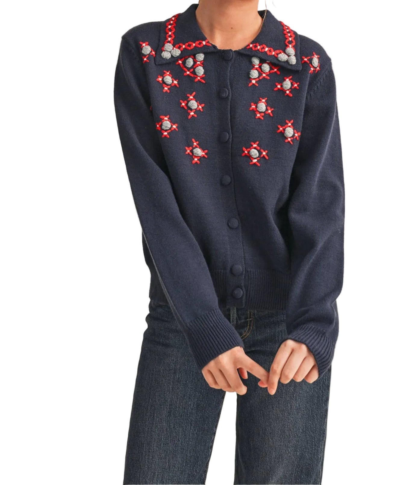 A woman wearing a navy blue knit cardigan with red and white embroidered star patterns on the chest, paired with dark blue jeans, against a solid beige background.