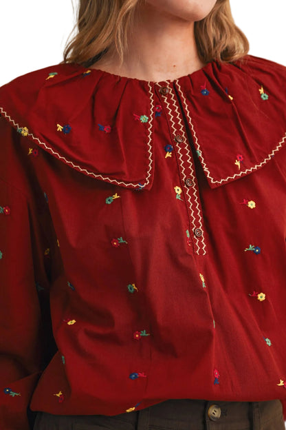 A close-up of a model wearing a red long-sleeve blouse with a large ruffled collar, white zigzag trim, and small colorful embroidered floral details.