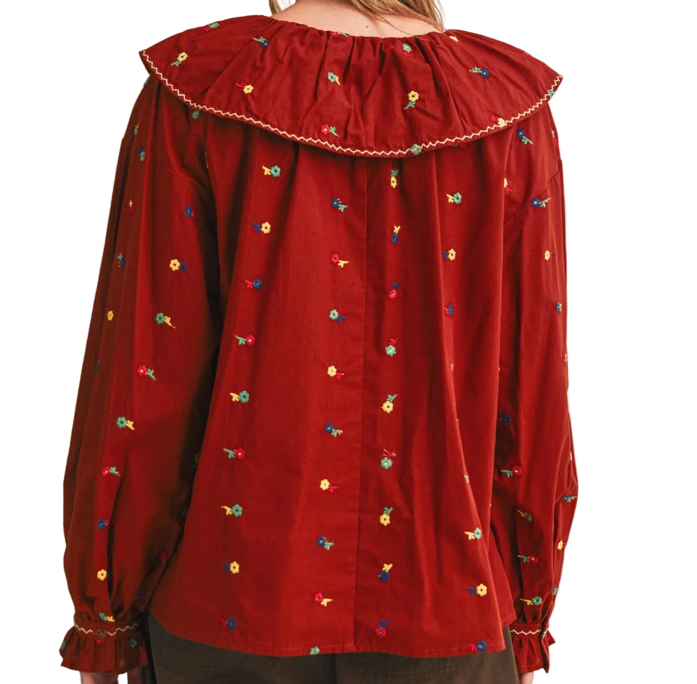 A woman shown from the back wearing a red long-sleeve blouse with a large ruffled collar and small multicolored embroidered motifs, paired with brown pants against a plain white studio background.