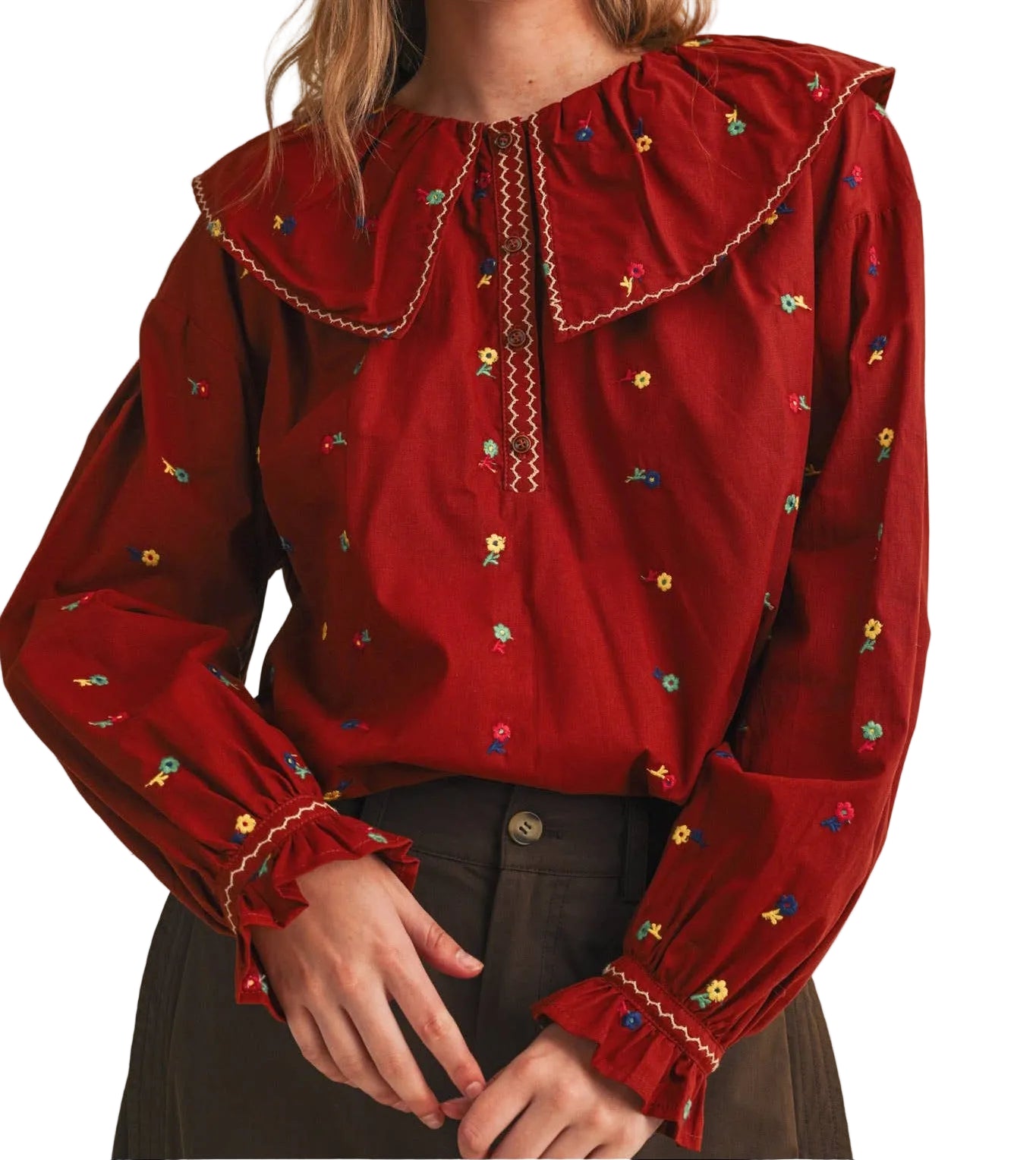 A woman wearing a deep red long-sleeve blouse with a large ruffled collar, embroidered details, and gathered cuffs, paired with dark brown trousers against a plain white  background.