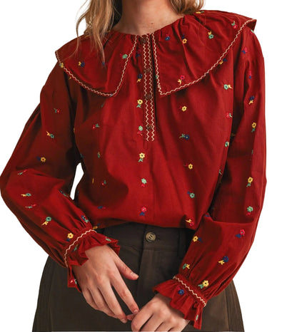 A woman wearing a deep red long-sleeve blouse with a large ruffled collar, embroidered details, and gathered cuffs, paired with dark brown trousers against a plain white  background.