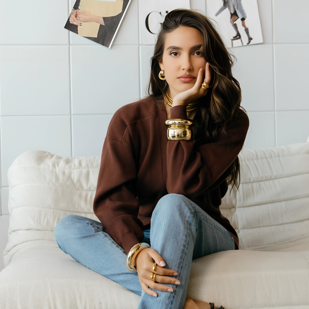 Woman sitting on a white couch wearing a brown sweater and blue jeans, with gold jewelry.