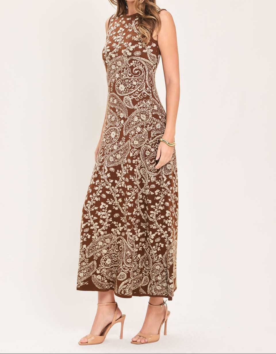 A woman modeling a sleeveless, high-neck maxi dress with an intricate brown and beige paisley pattern, paired with nude high-heeled sandals against a plain light background.