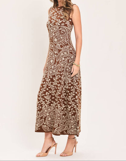 A woman modeling a sleeveless, high-neck maxi dress with an intricate brown and beige paisley pattern, paired with nude high-heeled sandals against a plain light background.